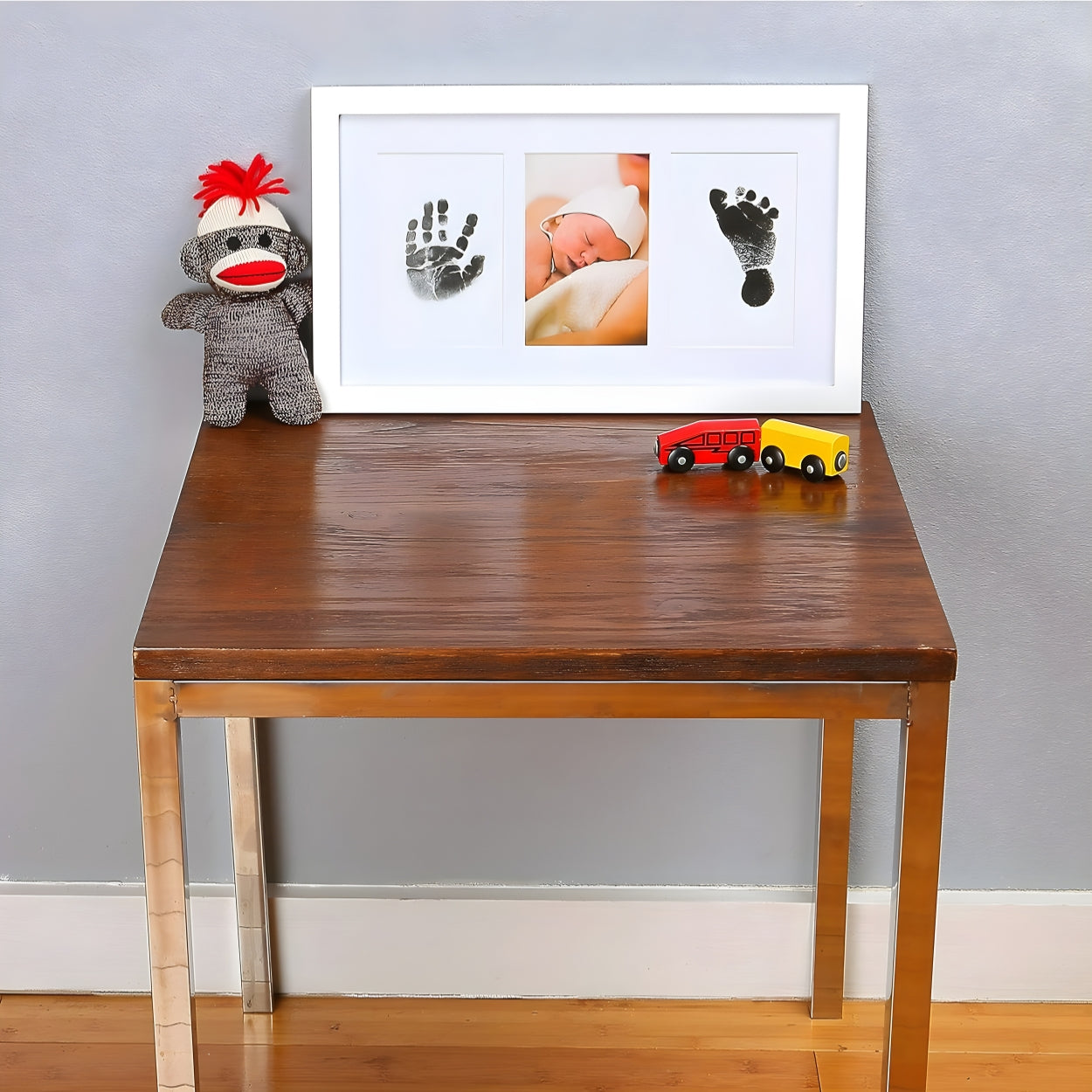 Wooden table with a framed baby photo and handprints, a sock monkey, and toy cars against a gray wall. - Little Treasures